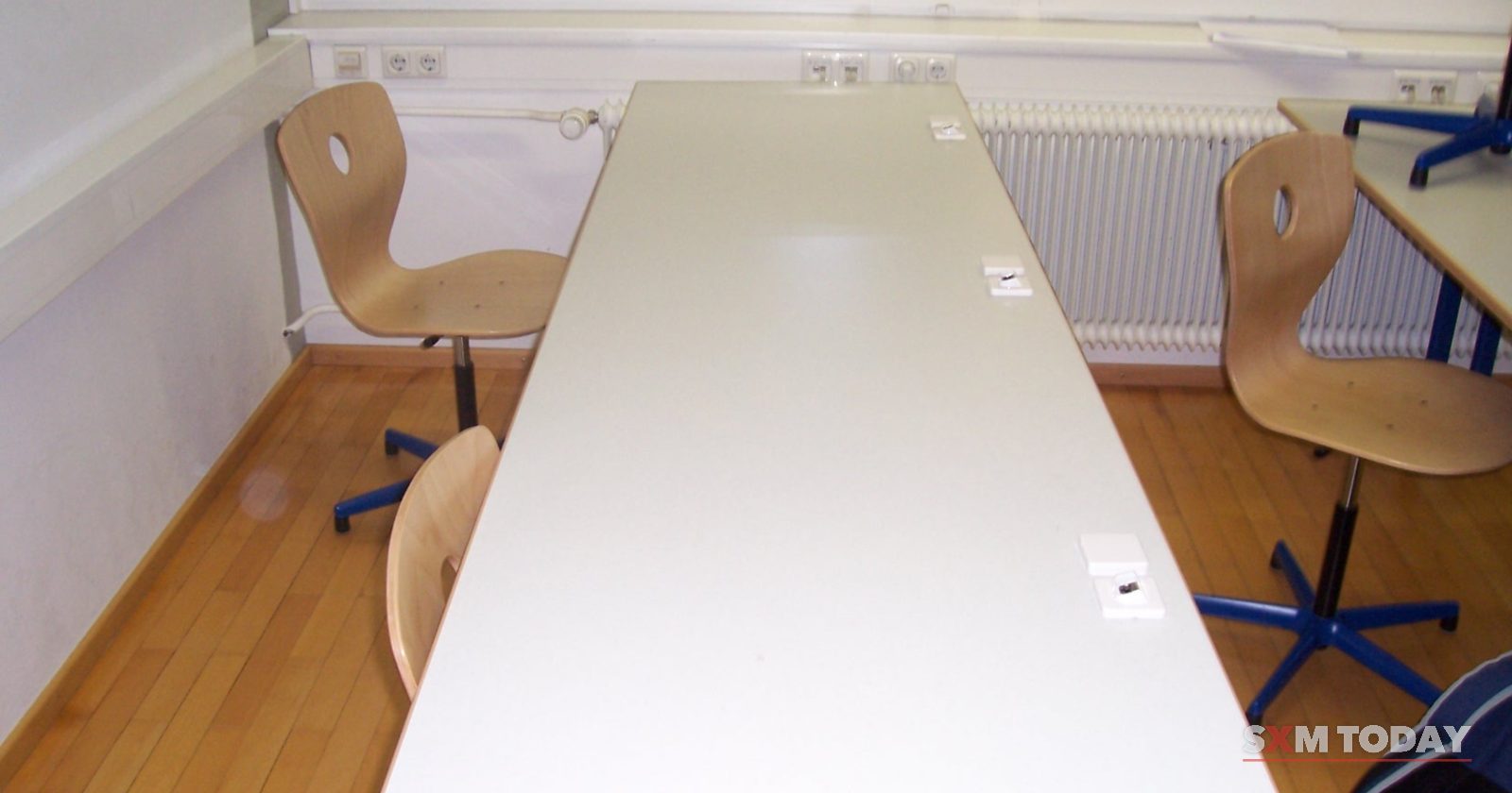 A long white desk with swivel chairs on either side in a bright, empty classroom with blinds on the windows.