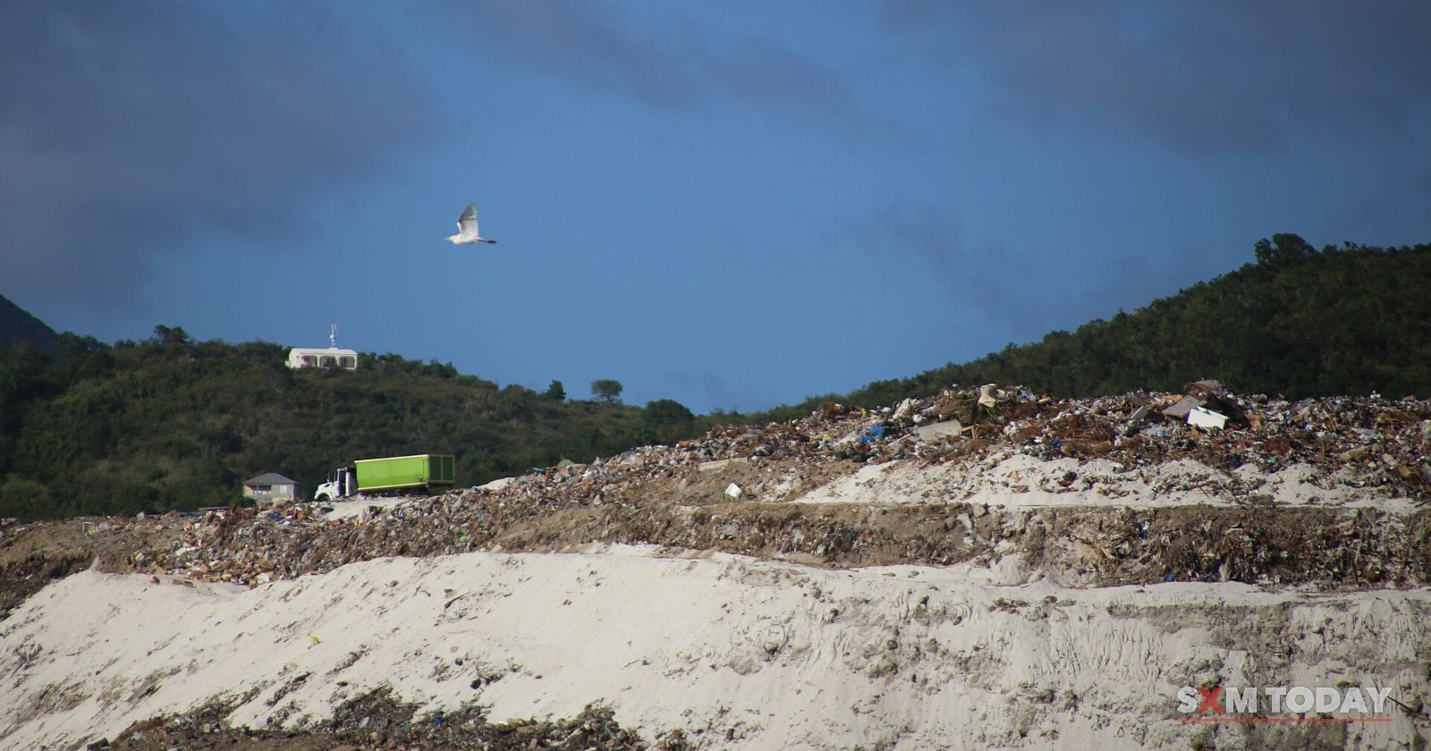 A mound of dumped waste and rubble stretches under a dark stormy sky, with a green hillside and a truck in the distance.