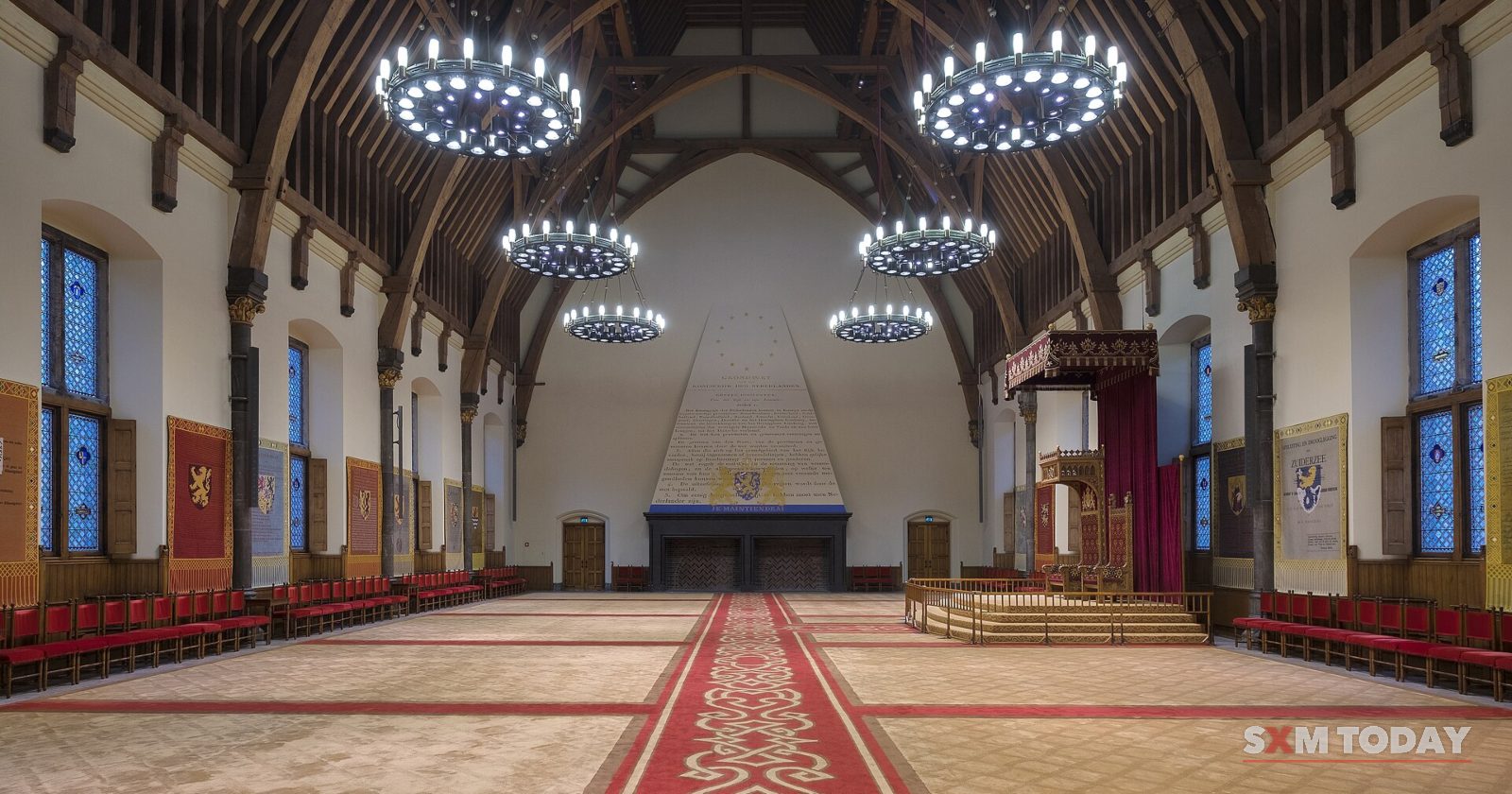 An empty great hall with a timber roof, round chandeliers, a long red carpet and a raised dais at the far end.