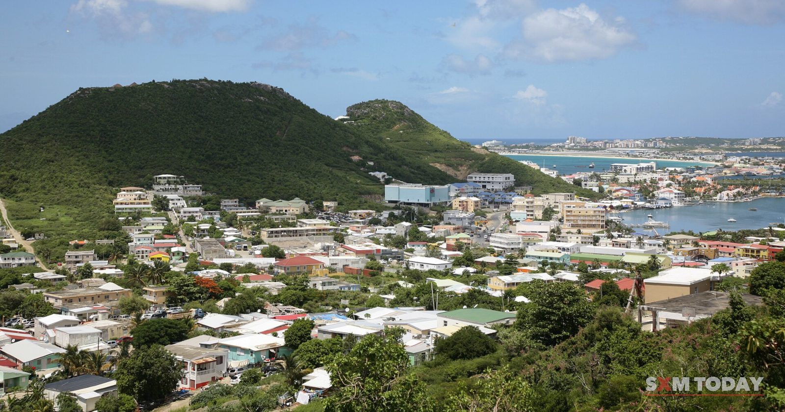Houses cover a hillside below a rounded green hill, with a lagoon and waterfront buildings beyond under a blue sky.