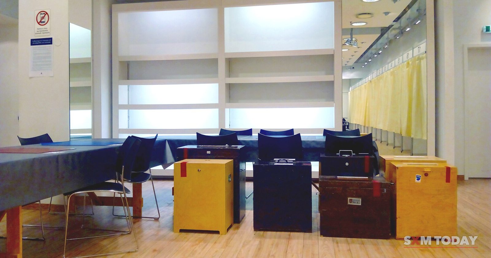 Wooden ballot boxes sit on a table in front of curtained voting booths in an empty room.