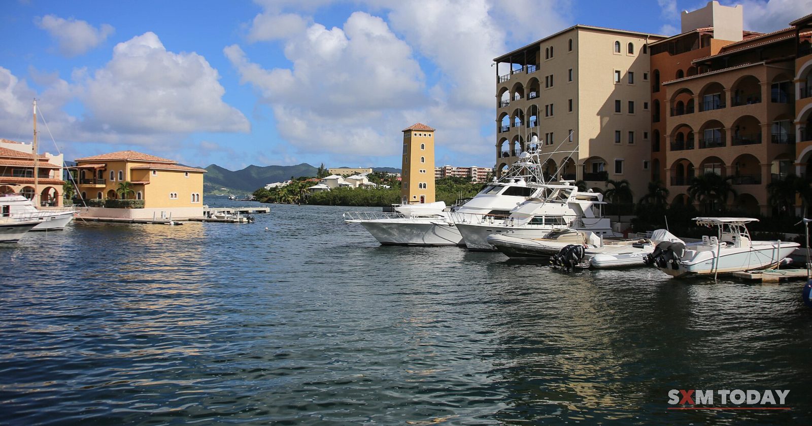 Motor boats are moored in a marina of dark blue water, ringed by terracotta and cream apartment buildings with a tower, under a blue sky with clouds.