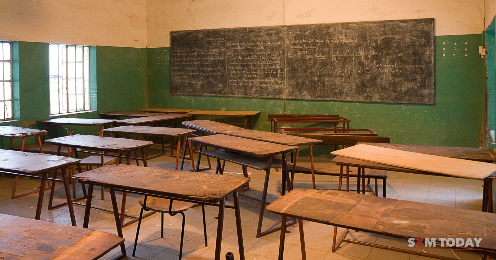 An empty classroom with rows of worn wooden desks and benches facing a chalkboard on a green-painted wall.
