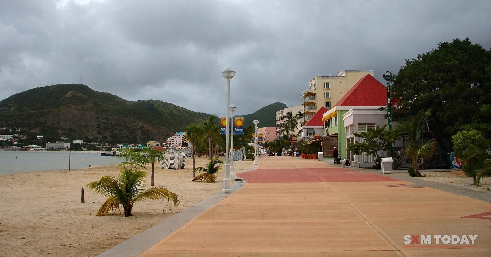 A paved boardwalk runs beside a sandy beach with young palms and low buildings under a cloudy sky.