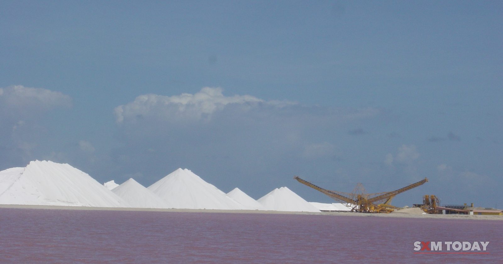 Piles of white salt and a conveyor stand behind a pink salt pond under a blue sky.
