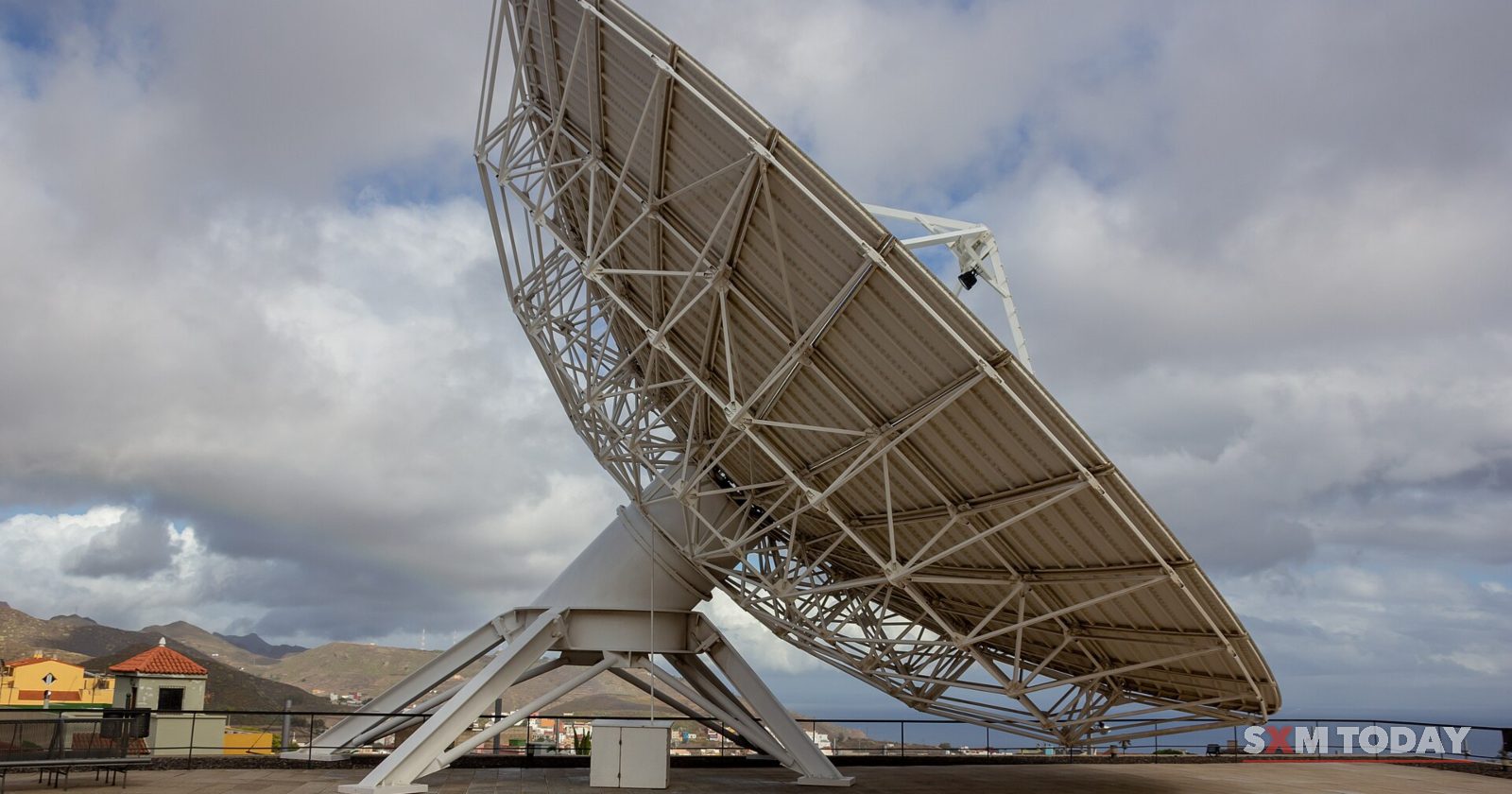 A large lattice-backed dish antenna tilted skyward on a paved terrace under broken cloud.