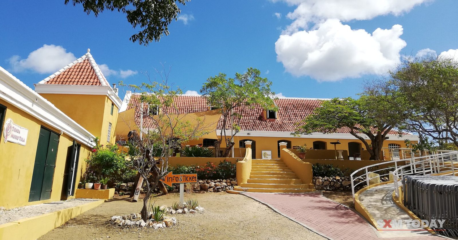 A yellow colonial building with a red-tiled roof and steps leading up to it, framed by trees under a blue sky.