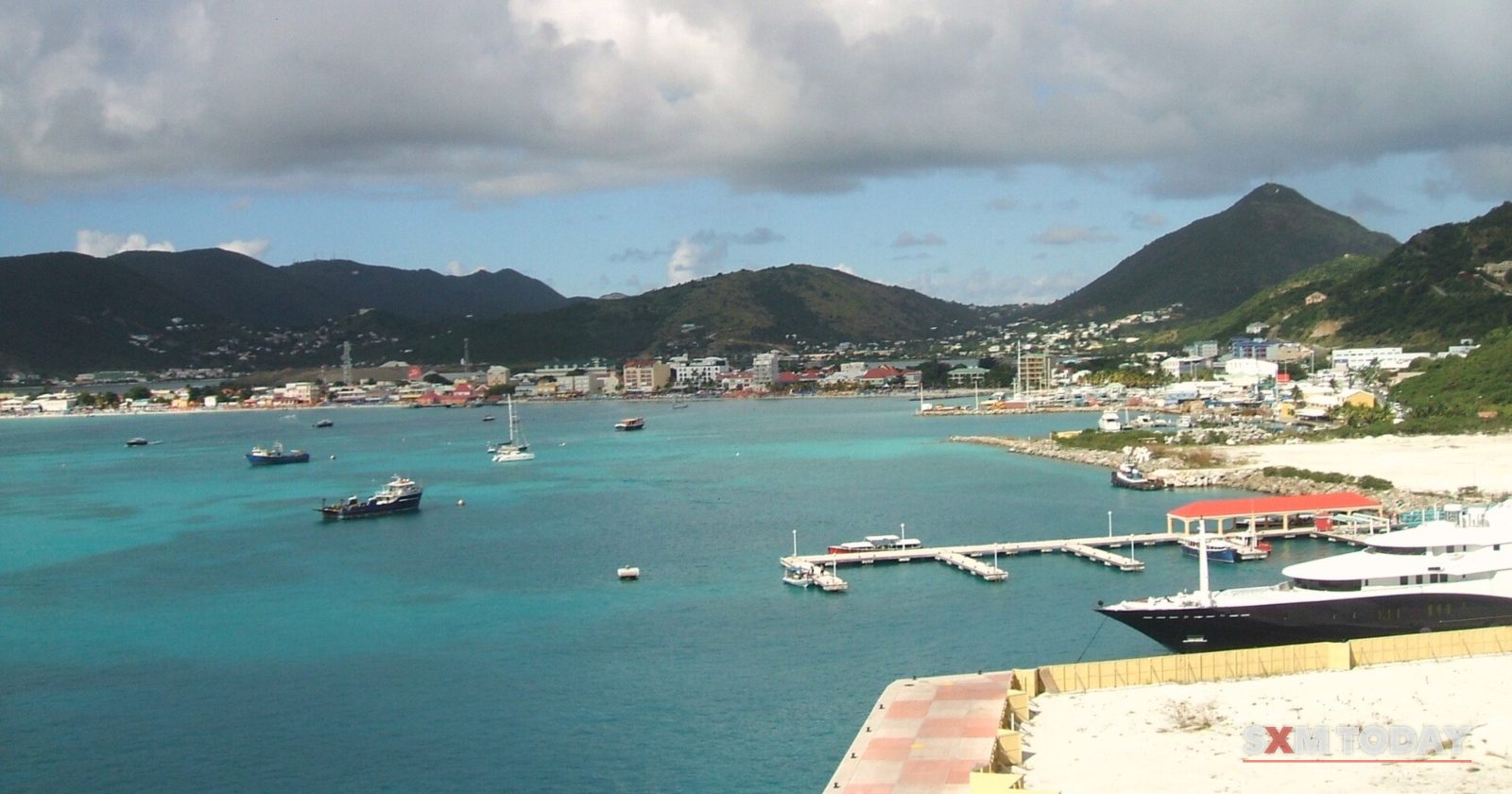 From a high vantage point, turquoise harbour water with a small pier and moored boats stretches toward the town and green hills, with a yacht in the foreground.