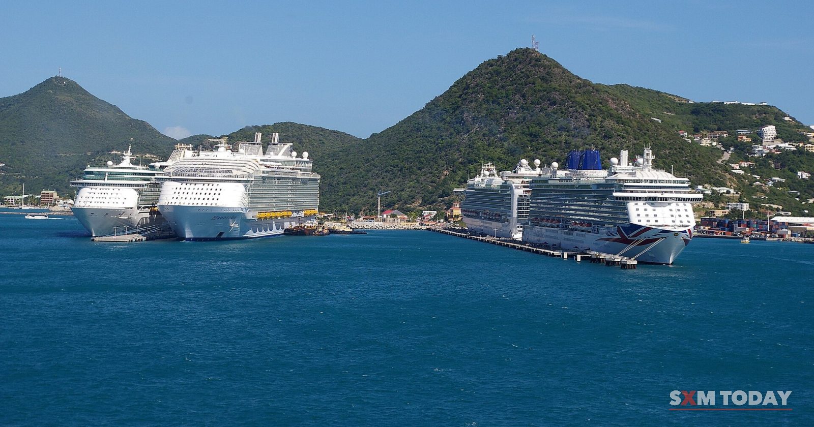 Four large cruise ships are docked in a blue bay below green hills.