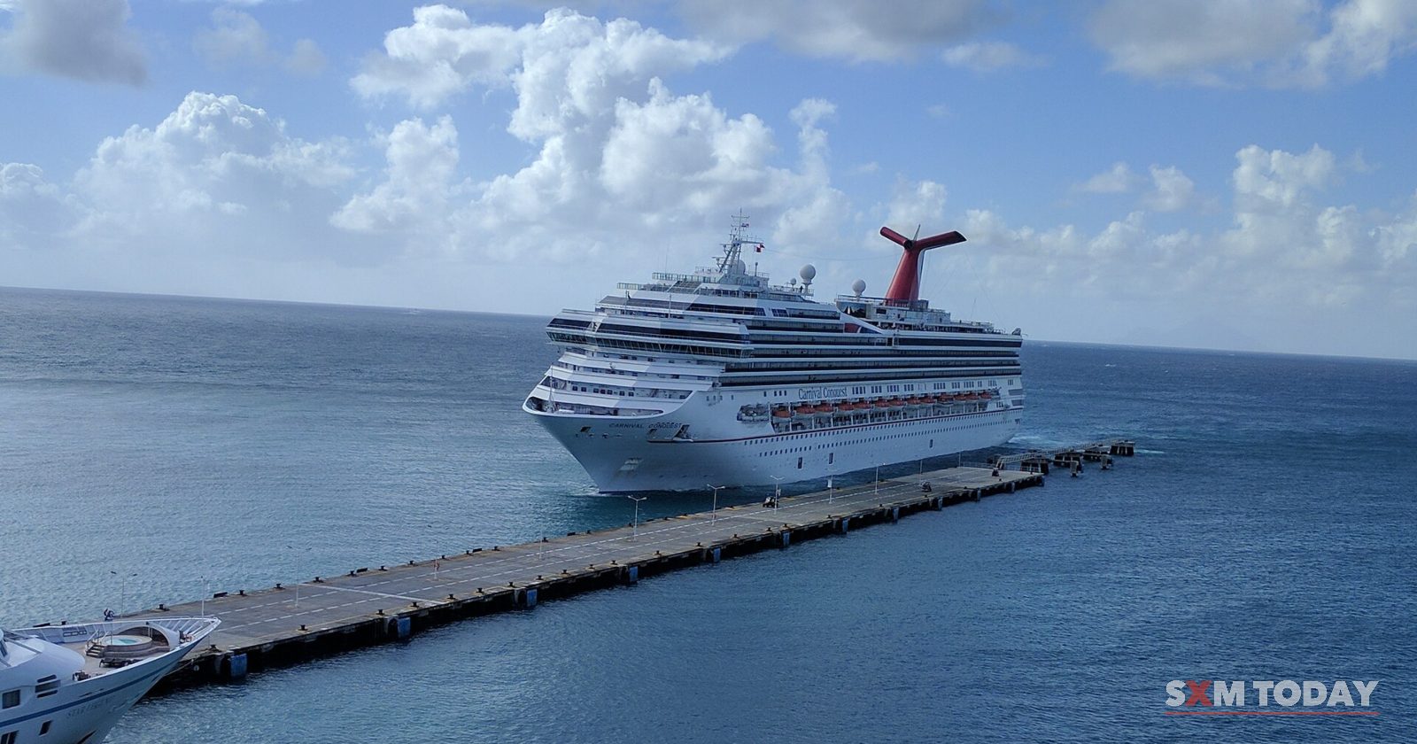 A large white cruise ship is moored at the end of a long concrete pier in calm blue water, under a partly cloudy sky.