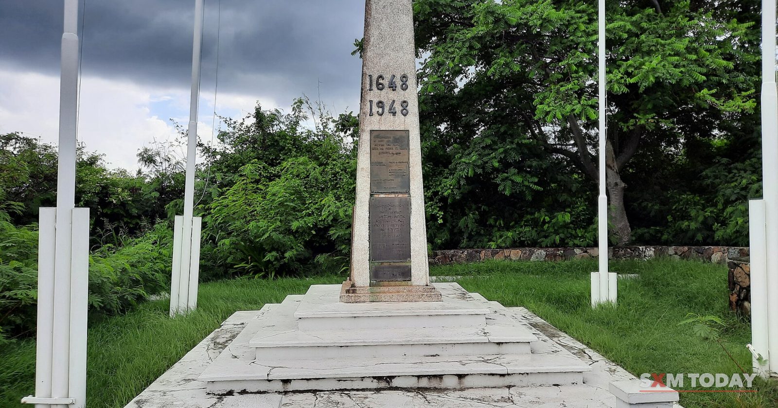 A white stone obelisk inscribed 1648 and 1948 stands on a stepped plinth between flagpoles, with trees and a cloudy sky behind.