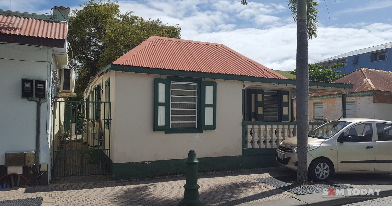 A single-storey cream house with a red roof and green shutters sits behind a low wall on a street corner, with a small car parked in front.