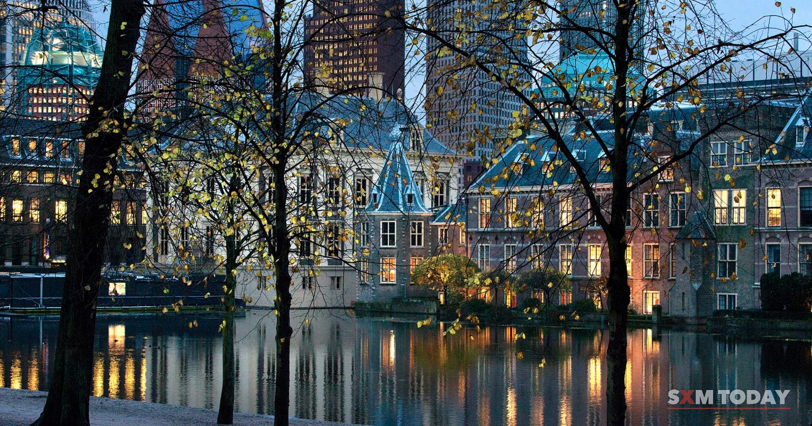 A small corner tower and brick buildings lit at dusk and reflected in a pond, with bare trees and office towers behind.