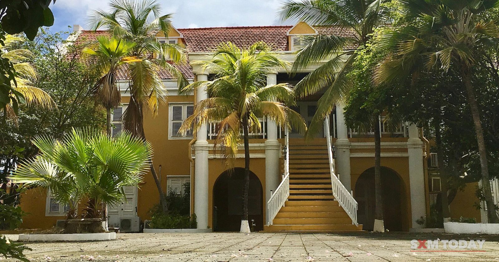 A two-storey yellow colonial government building with a central staircase, framed by palm trees under a blue sky.