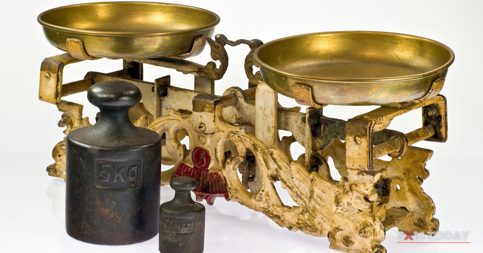 An ornate brass two-pan balance scale with two iron weights beside it on a white background.