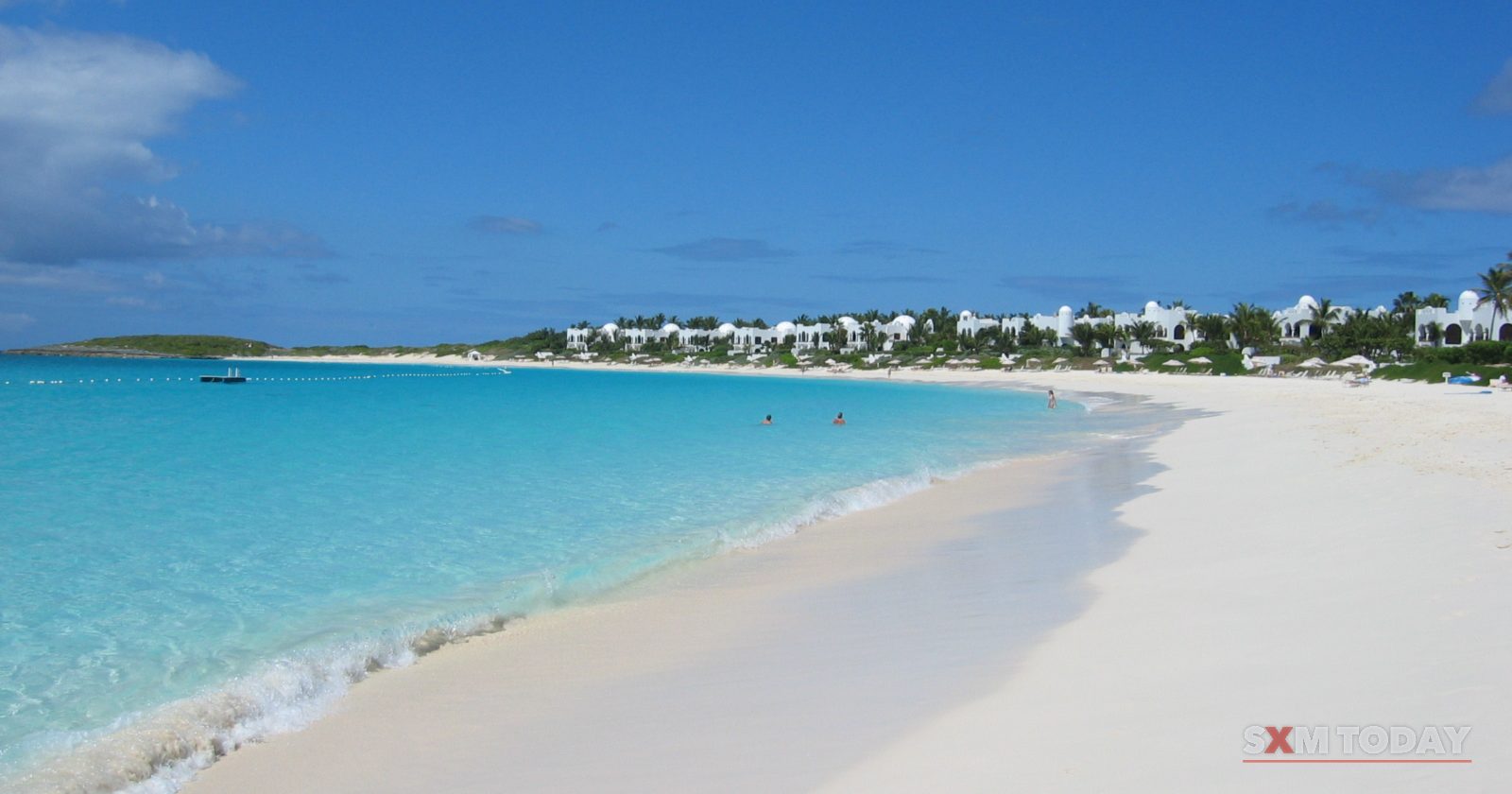 A wide white sand beach curves along clear turquoise water, with low white buildings along the far shore under a deep blue sky.