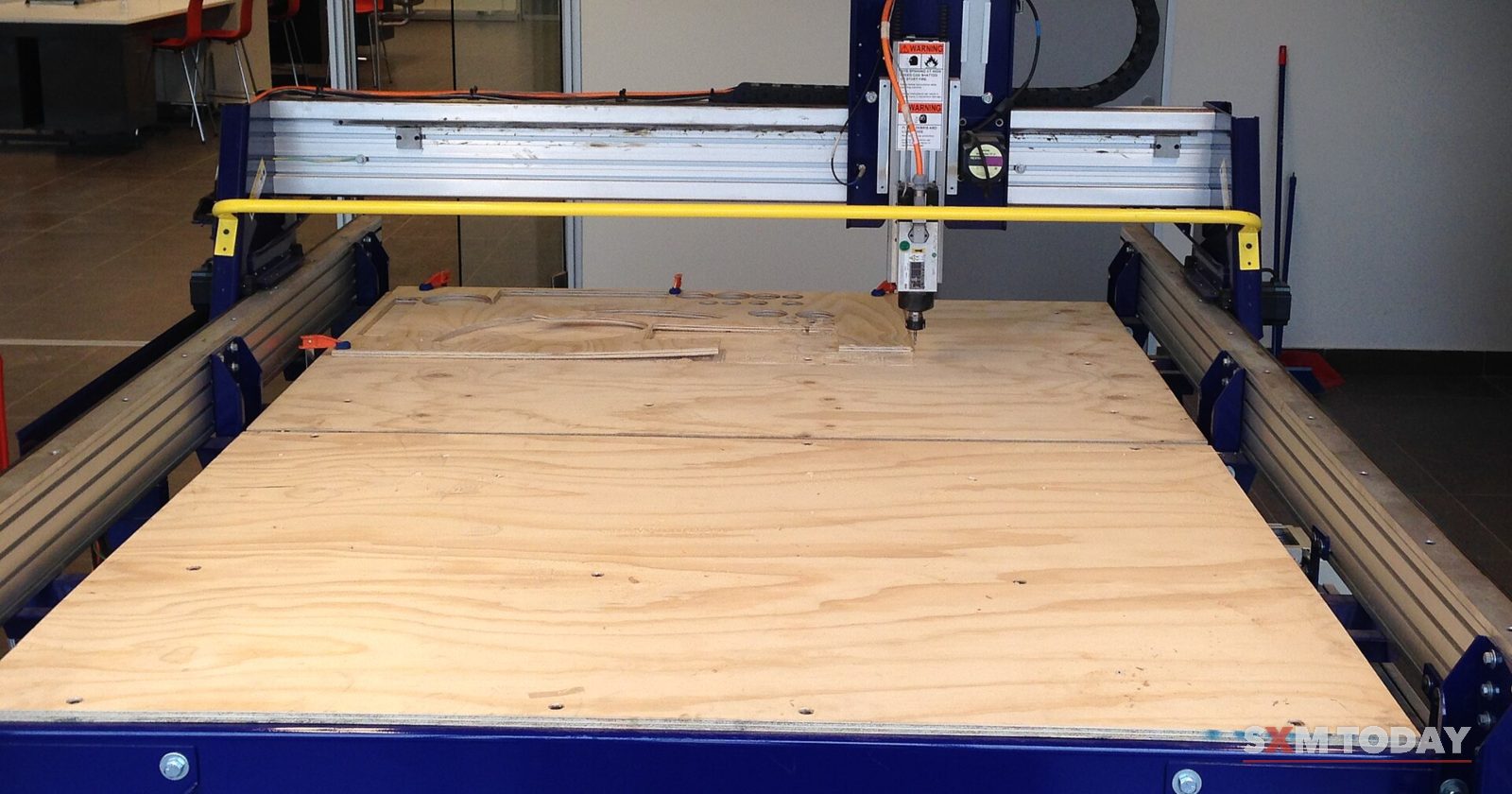 A large computer-controlled cutting machine with a plywood bed stands in an empty workshop.