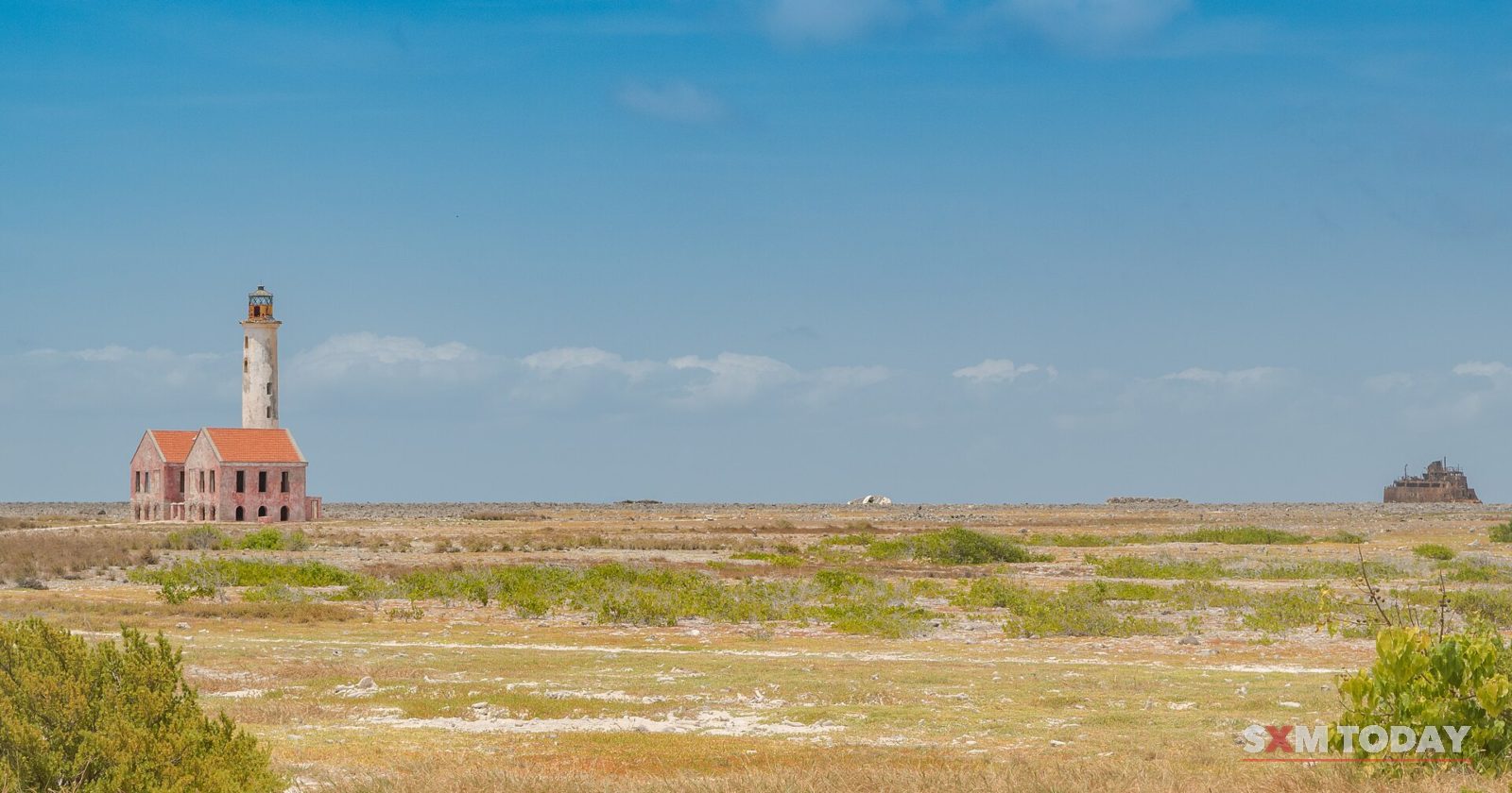 A white lighthouse and a pink-roofed keeper's house stand alone on a flat plain of dry grass under a blue sky.