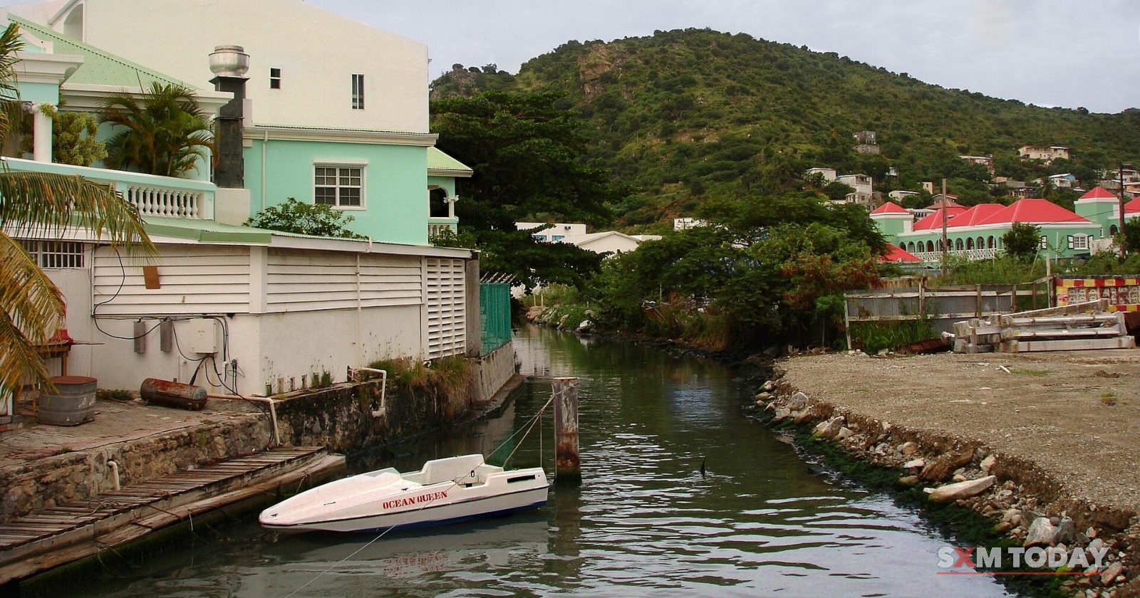 A narrow channel with a small speedboat runs between a stone bank and pastel houses, with green hills under a grey sky.