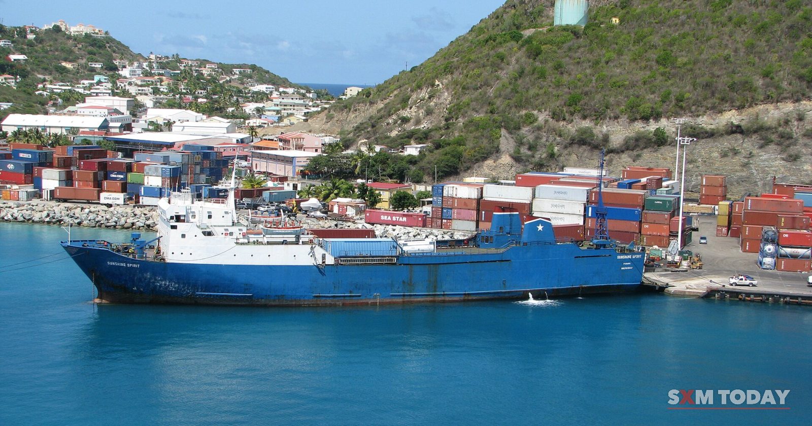 A blue cargo ship lies in turquoise water beside a container terminal stacked with shipping containers, below a green hillside.