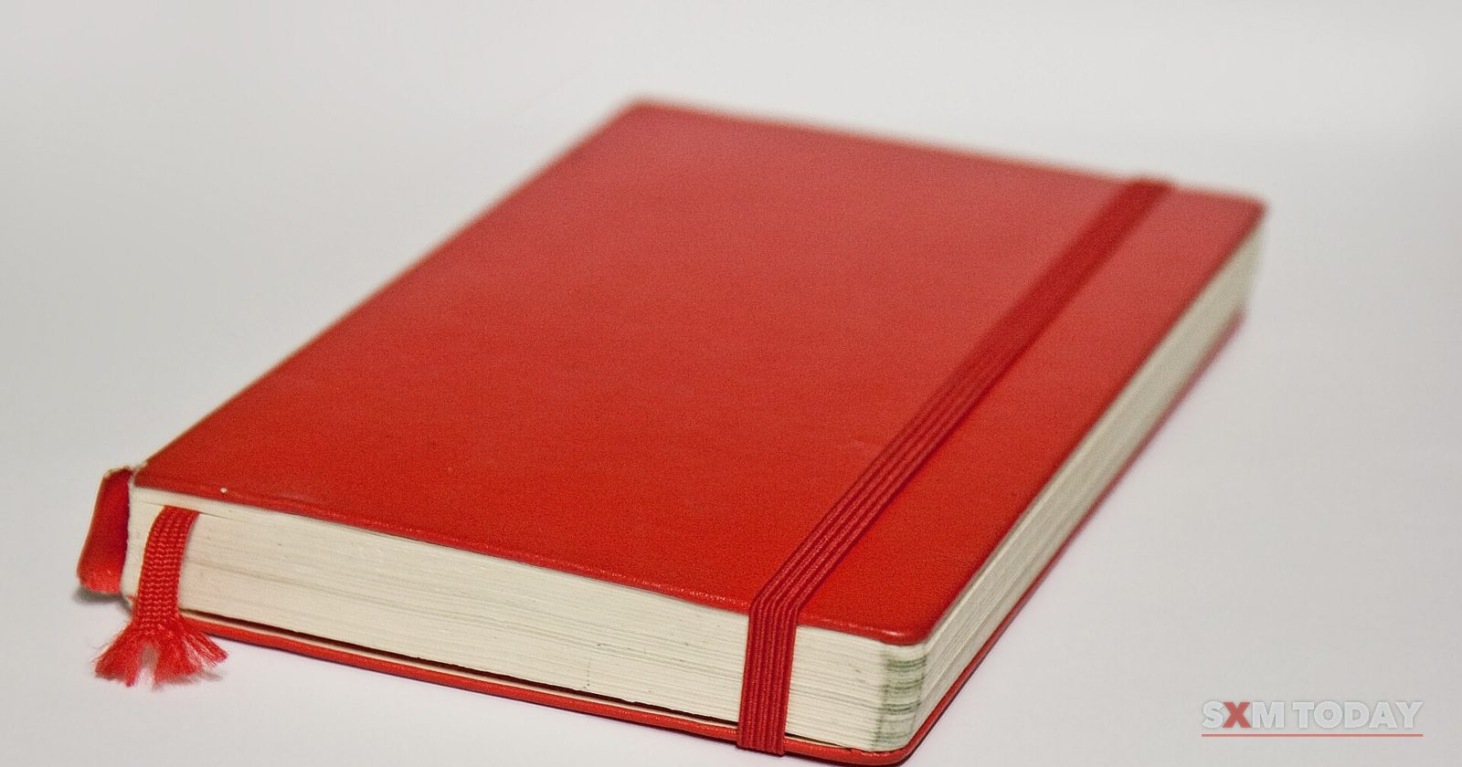 A red hardback notebook with an elastic band and a ribbon bookmark lies on a white surface.