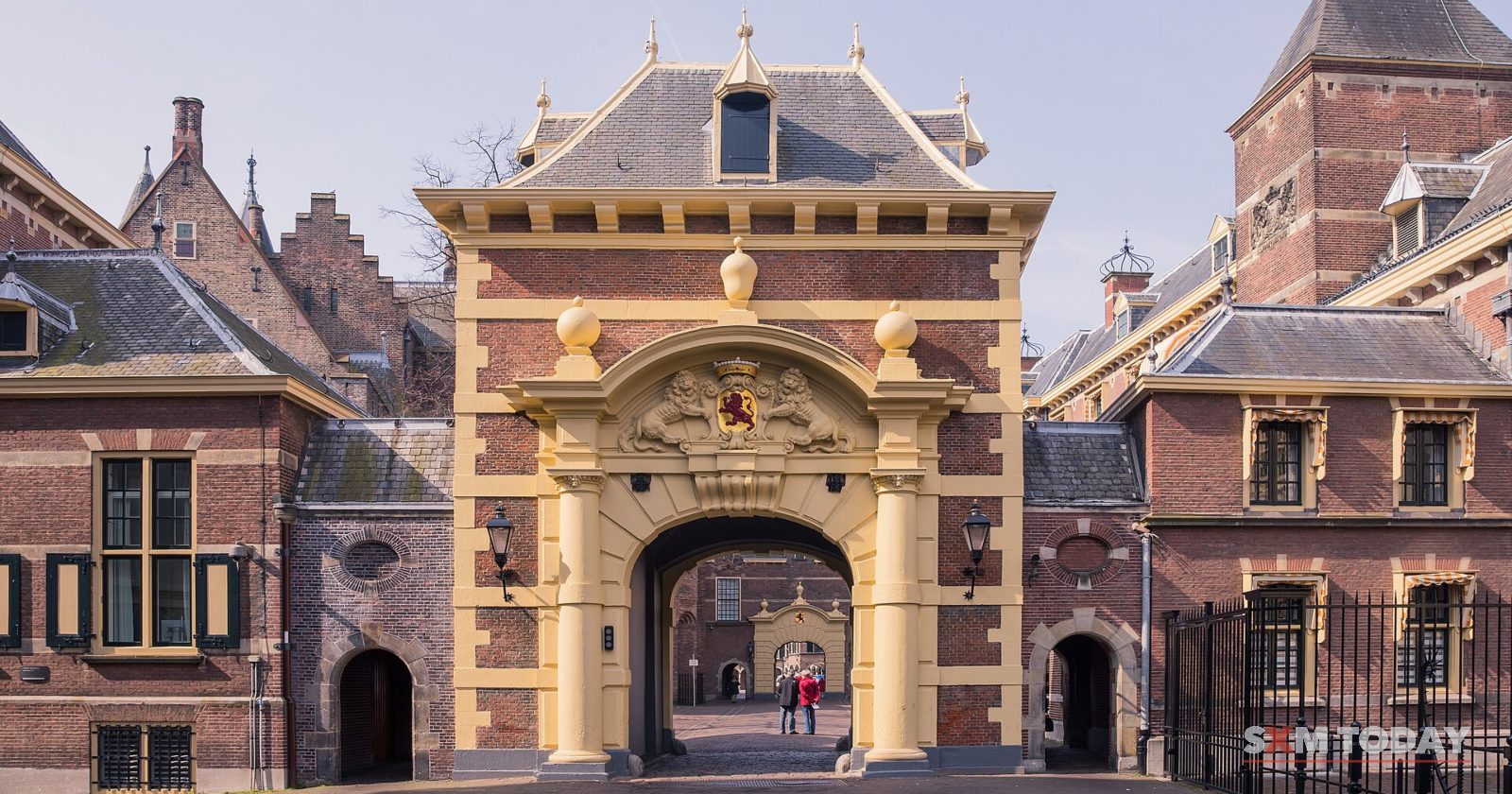 An ornate sandstone gateway with a coat of arms set between brick buildings, a cobbled square in front.