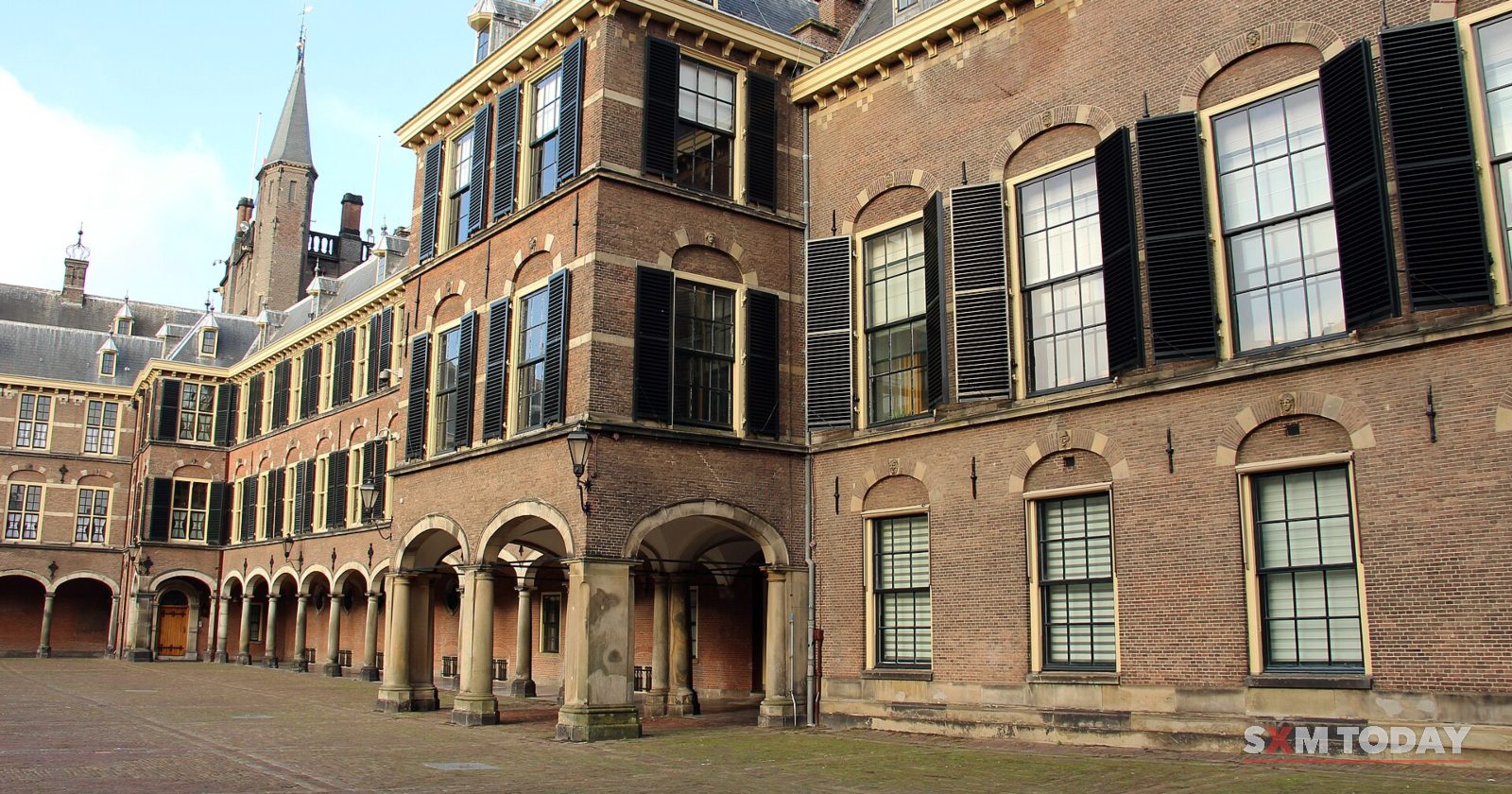 Brick buildings with dark shutters and an arcade on a sunlit cobbled courtyard, a spire behind.