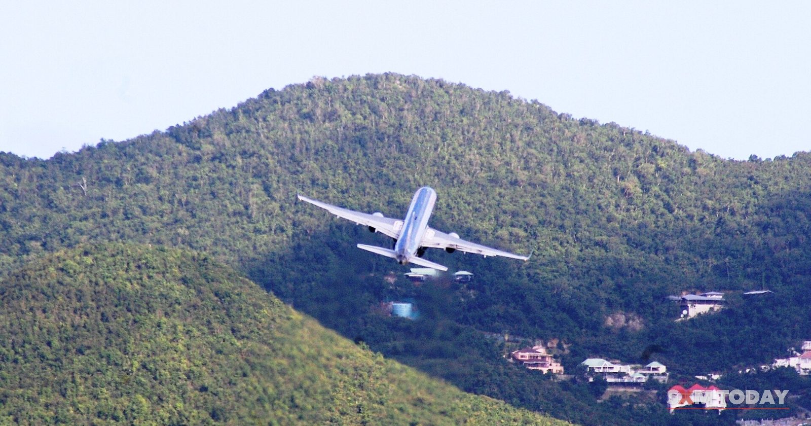 A white passenger jet climbs steeply over green hills dotted with houses.