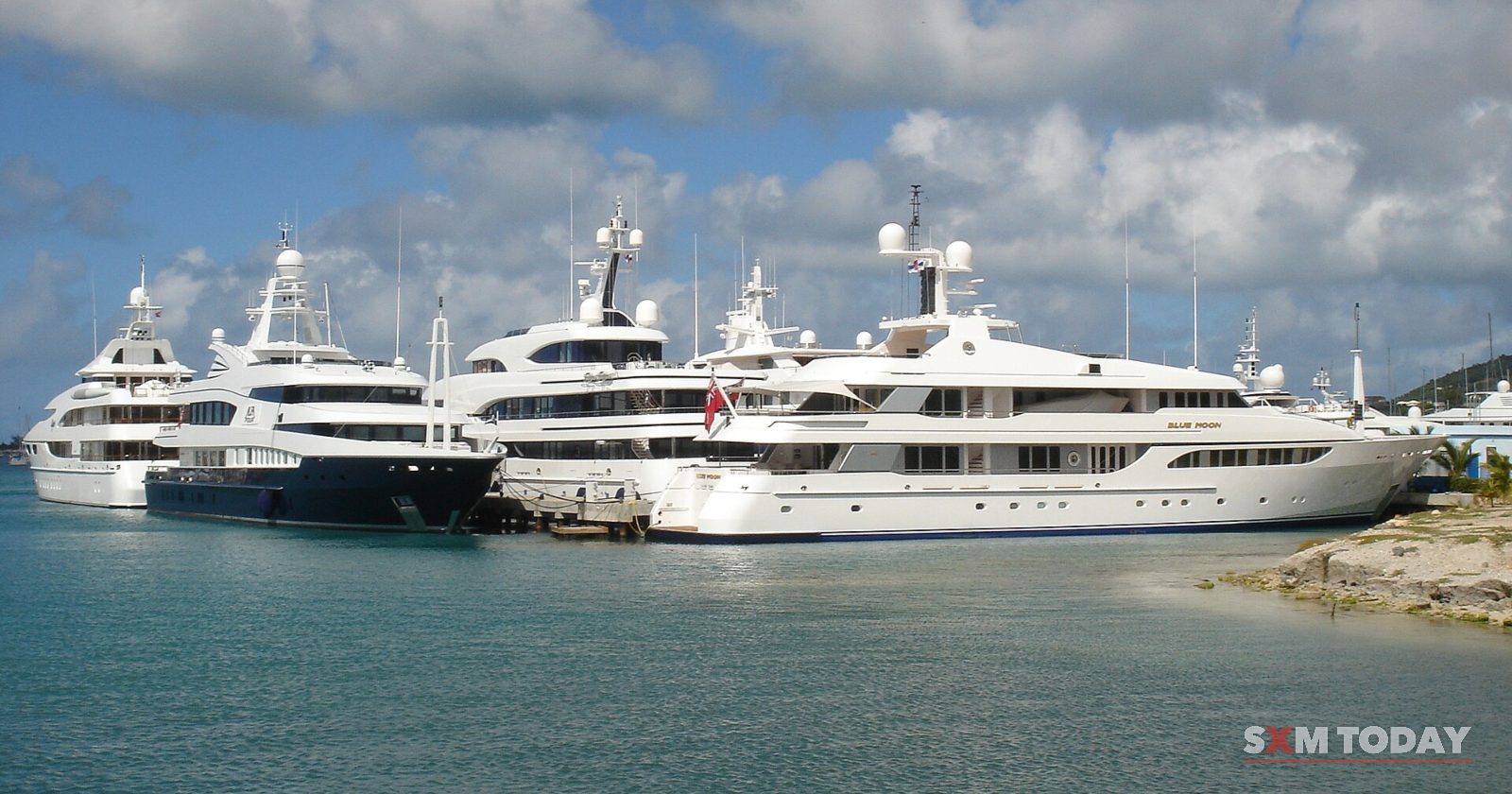 Several large white motor yachts are moored side by side in a marina under a partly cloudy sky.