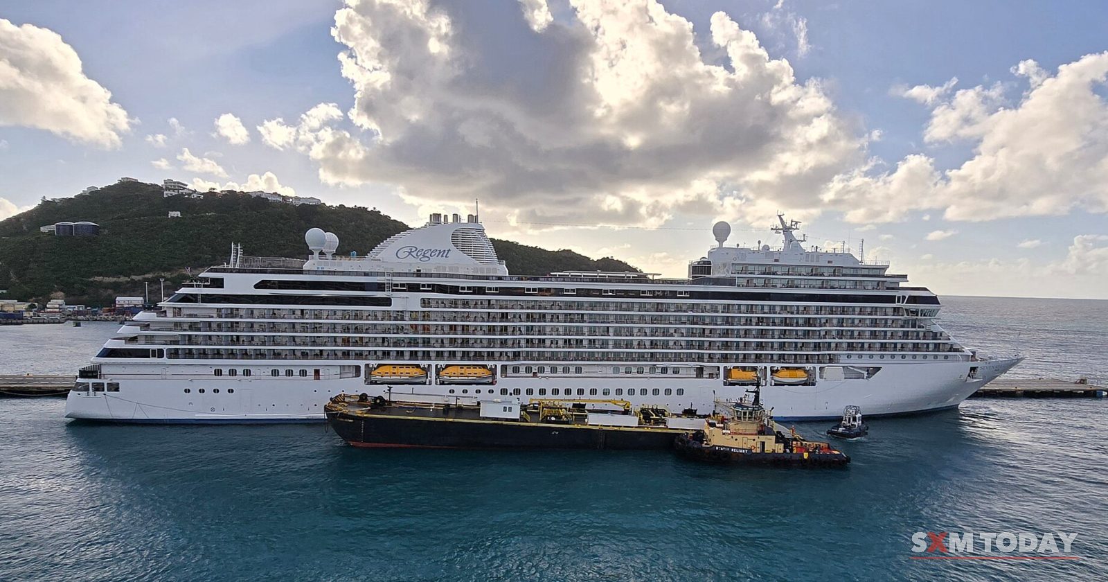 A large white cruise ship lies alongside a pier under a cloudy blue sky, with a smaller service vessel beside it and a green hill behind.