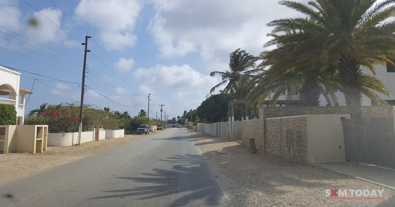 A quiet paved street lined with low walls, houses, palm trees and utility poles under a partly cloudy sky.