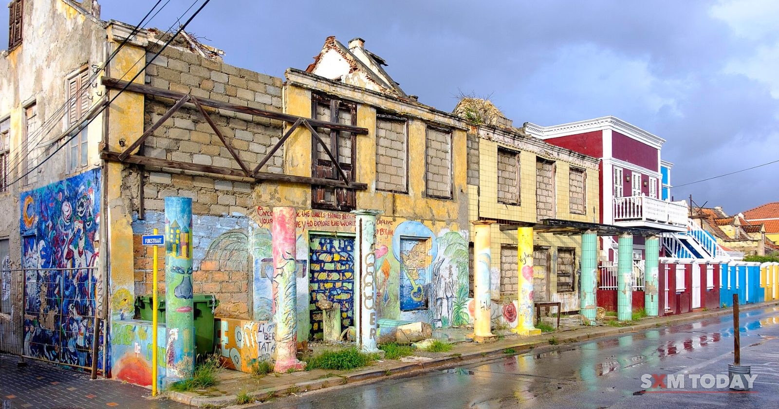 The roofless shell of an old two-storey building, its lower walls covered in colourful painted panels, stands on a rain-wet street.