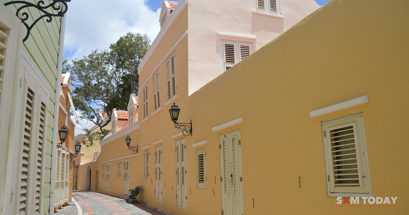 A narrow paved lane runs between restored yellow and pale-green colonial buildings with shuttered windows and wall lanterns.