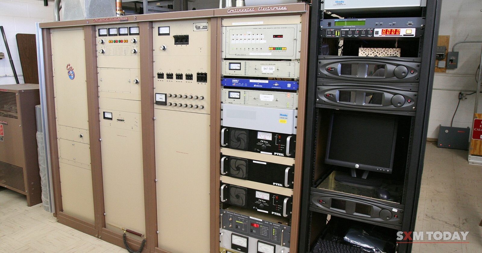 Beige transmitter cabinets and racks of broadcast equipment stand along the wall of an equipment room.