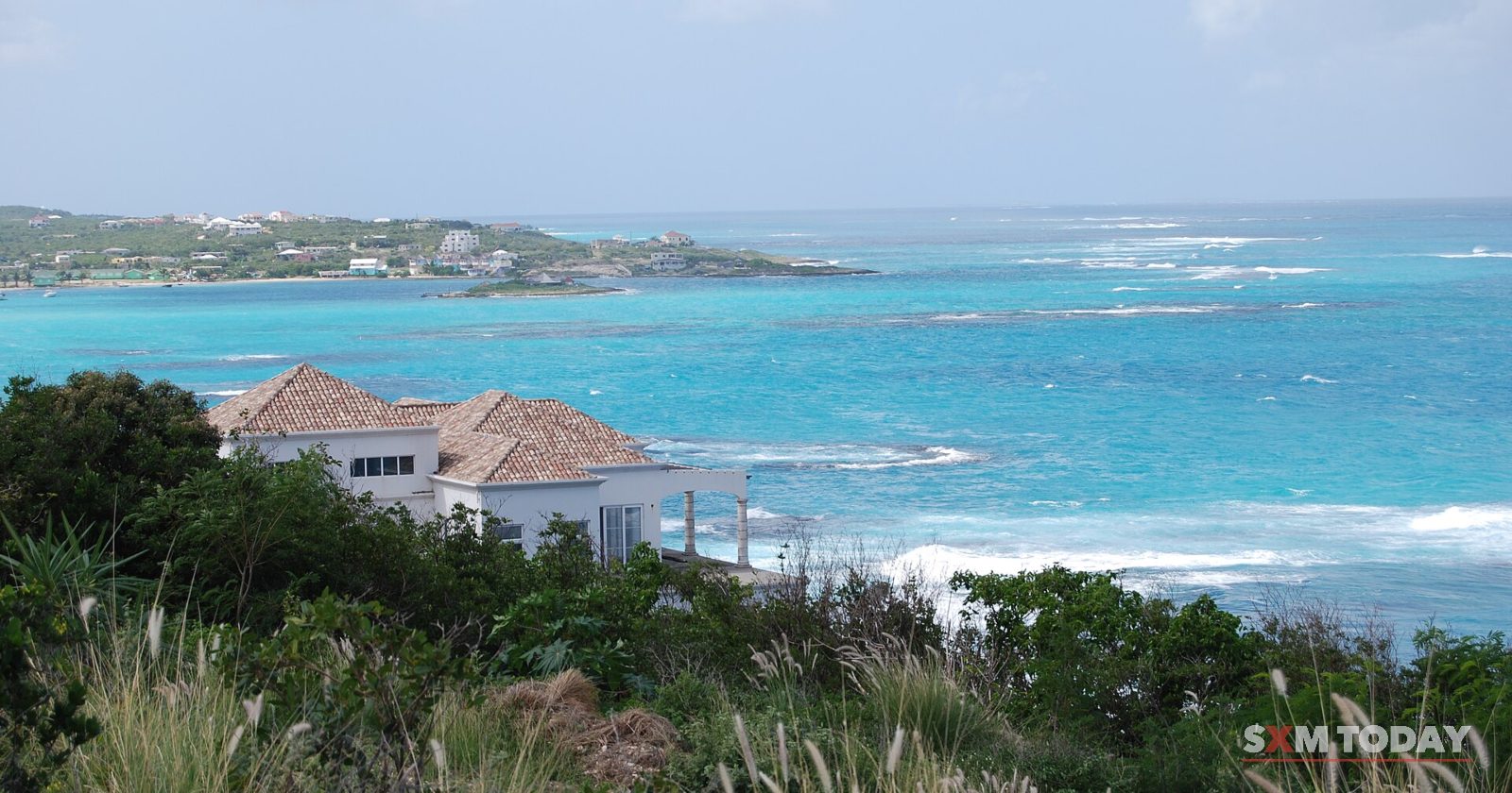 A view over coastal scrub and a red-roofed white house to a turquoise bay with breaking reef and a low headland beyond.