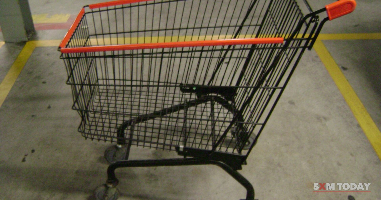 An empty shopping cart with orange handles standing on a concrete floor between yellow painted lines.