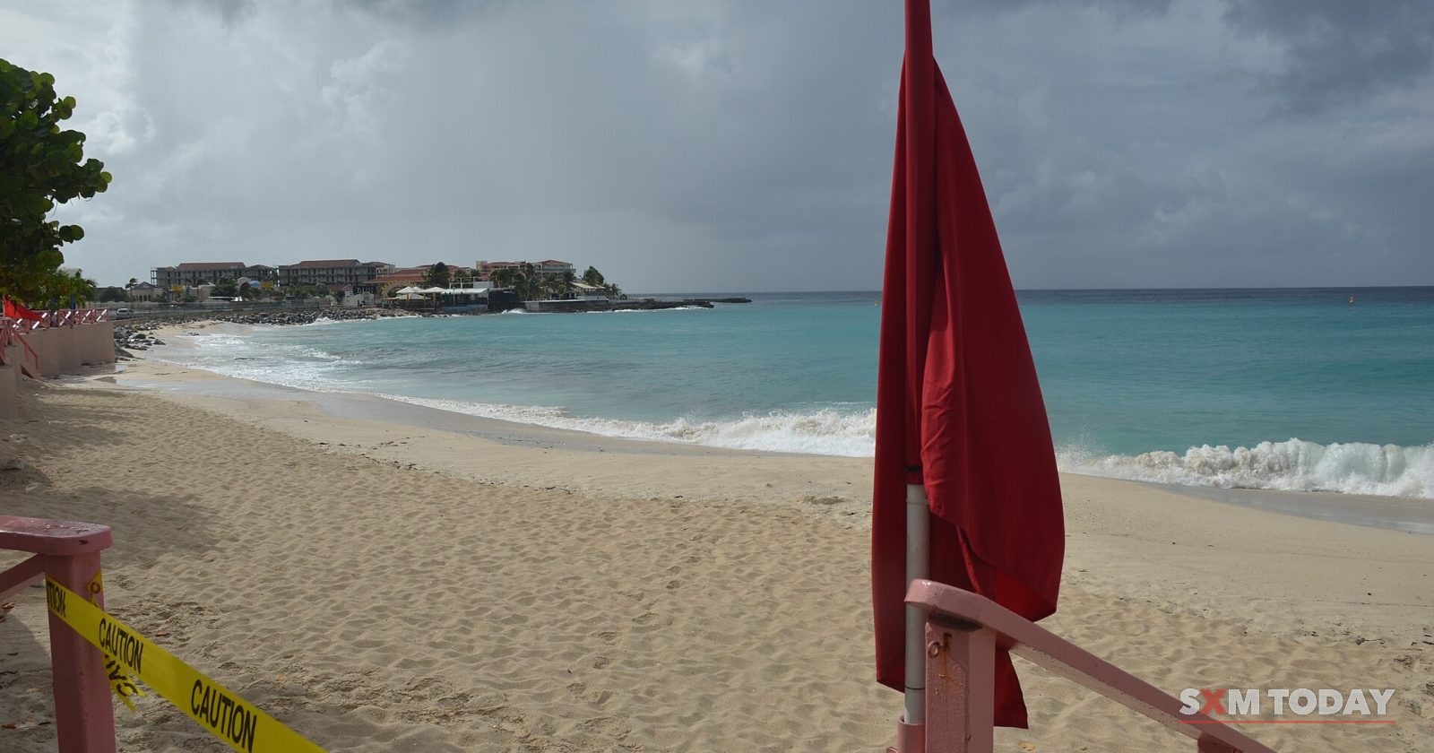 A red flag flies beside yellow caution tape on an empty beach as waves roll in under grey clouds.