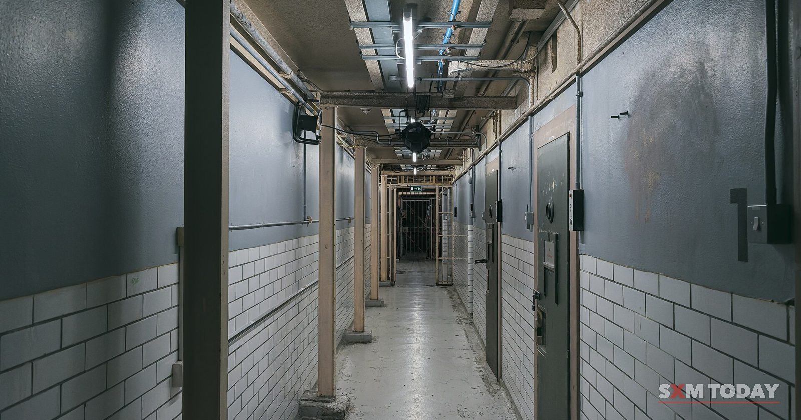 A narrow tiled corridor lined with heavy cell doors under ceiling pipes and a strip light.