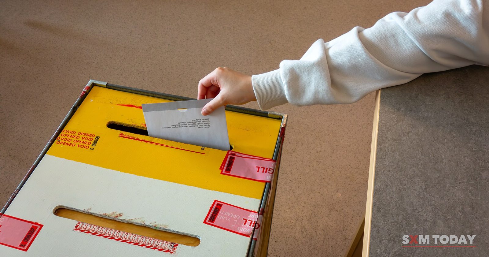 A hand drops an envelope into the slot of a sealed ballot box.