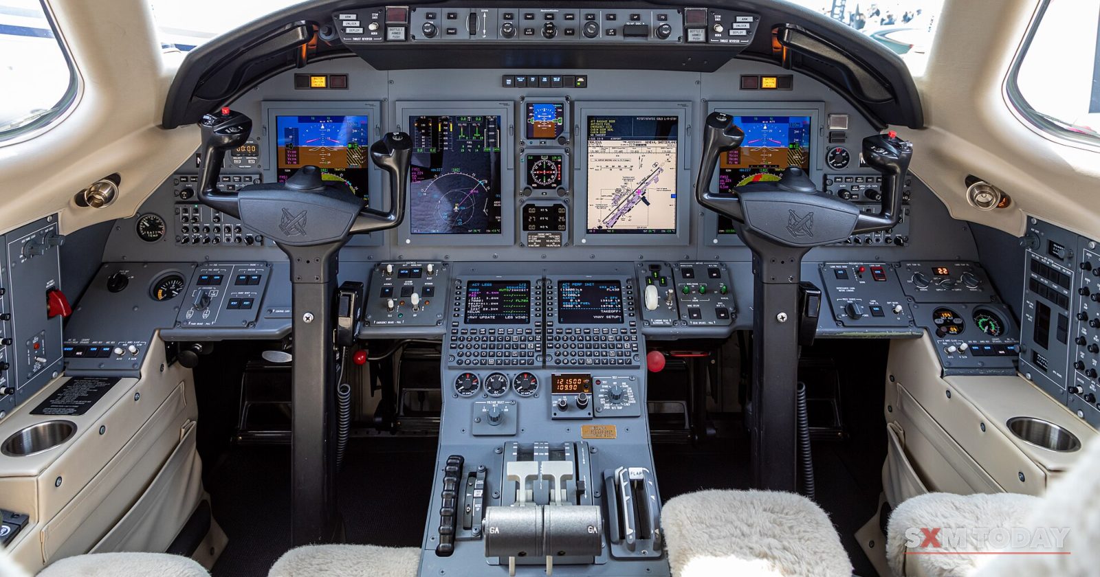 The empty cockpit of a small jet, with twin control yokes, glass instrument displays and throttle levers.