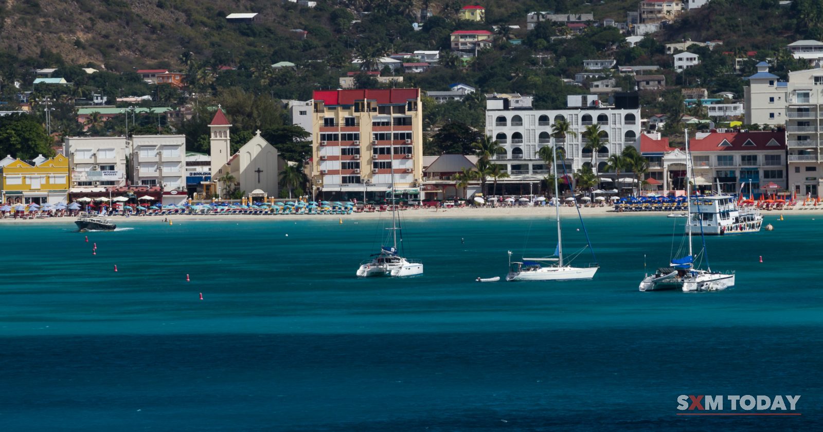 Sailboats are anchored in blue water in front of a beachfront lined with buildings, backed by hillsides dotted with houses.