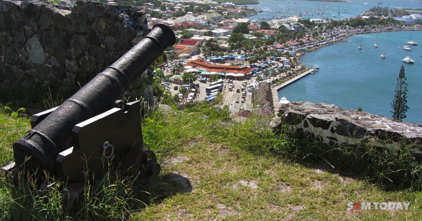 An old iron cannon on a hilltop points over red rooftops toward a blue bay and distant hills.