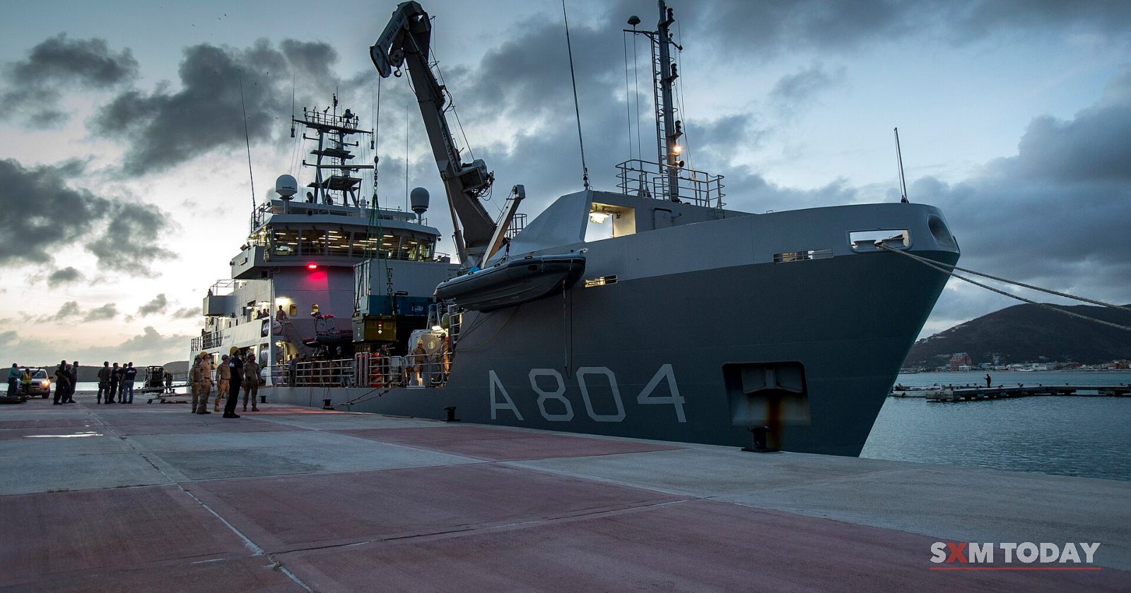 A grey naval support ship marked A804 is moored at a concrete pier under a dark cloudy sky, with small figures on the quay.