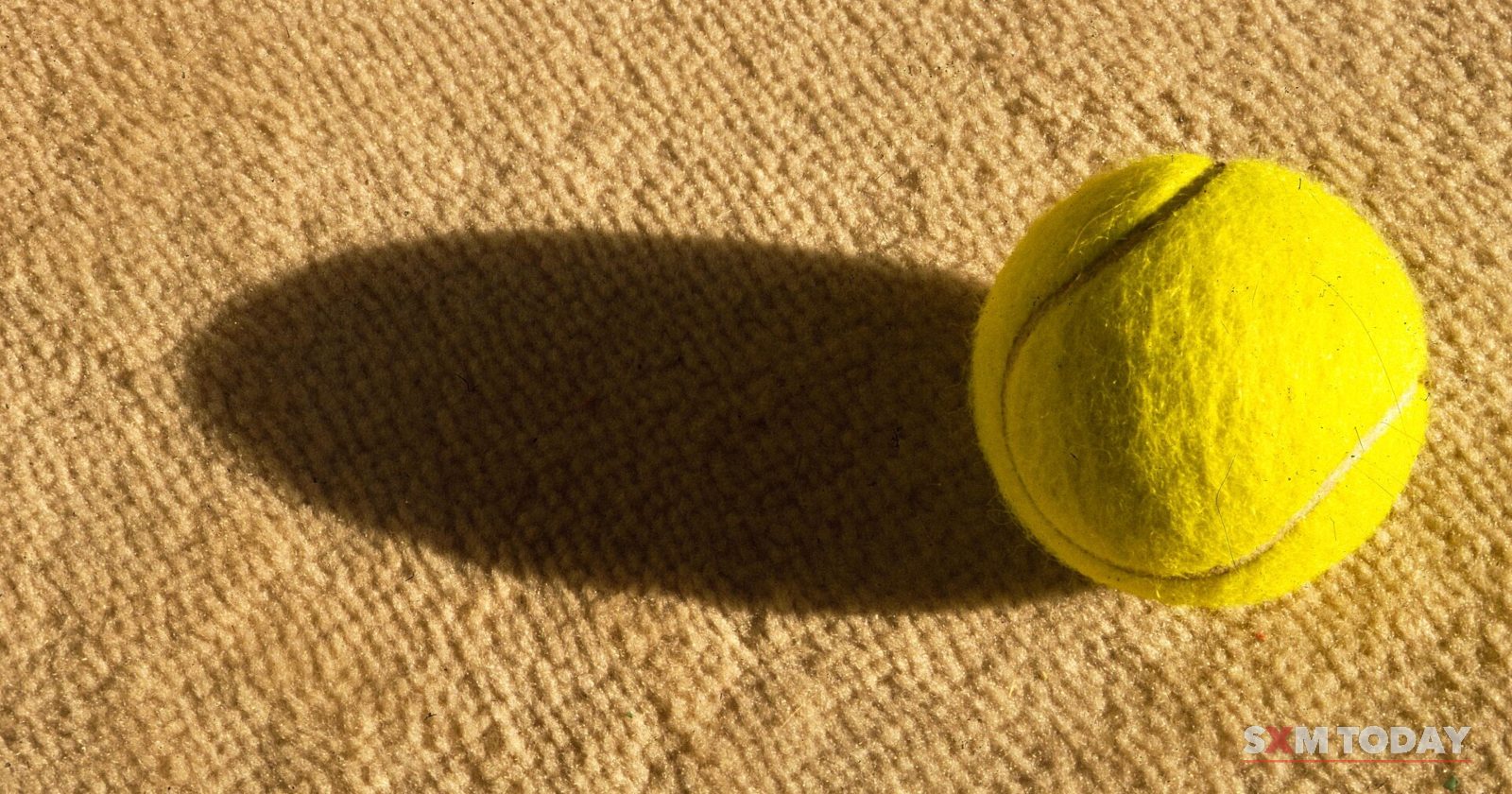 A single yellow tennis ball casts a long shadow across a sandy-coloured court surface.
