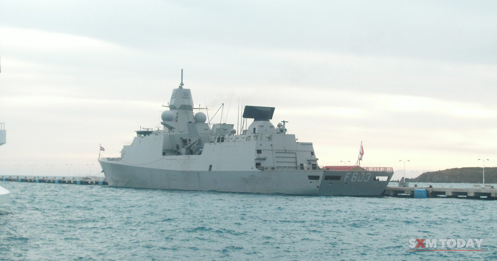 A grey naval frigate lies at anchor on choppy water under an overcast sky, with a low pier on the horizon.