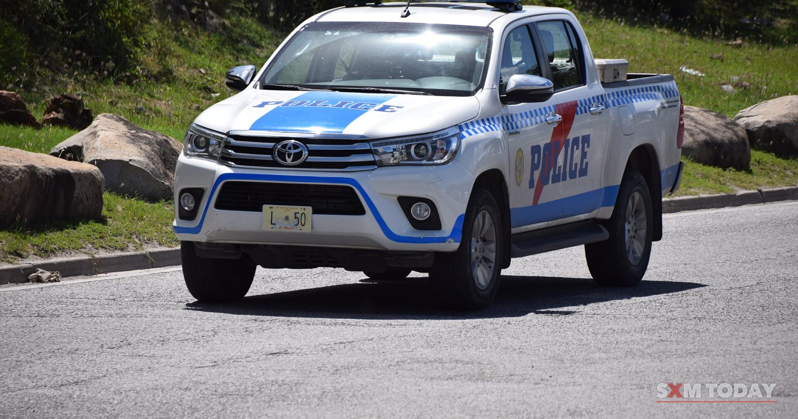 A white police pickup truck with blue and red markings drives on a road below a green hillside.