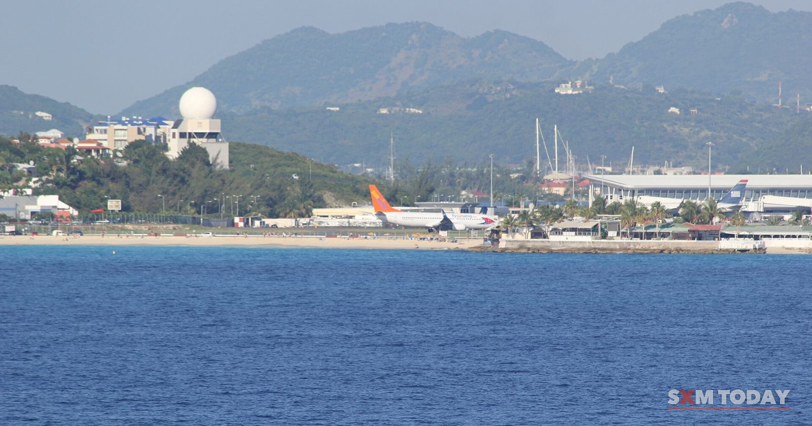 An airport terminal, parked jets and a white radar dome along the shoreline, with hills behind and open water in front.