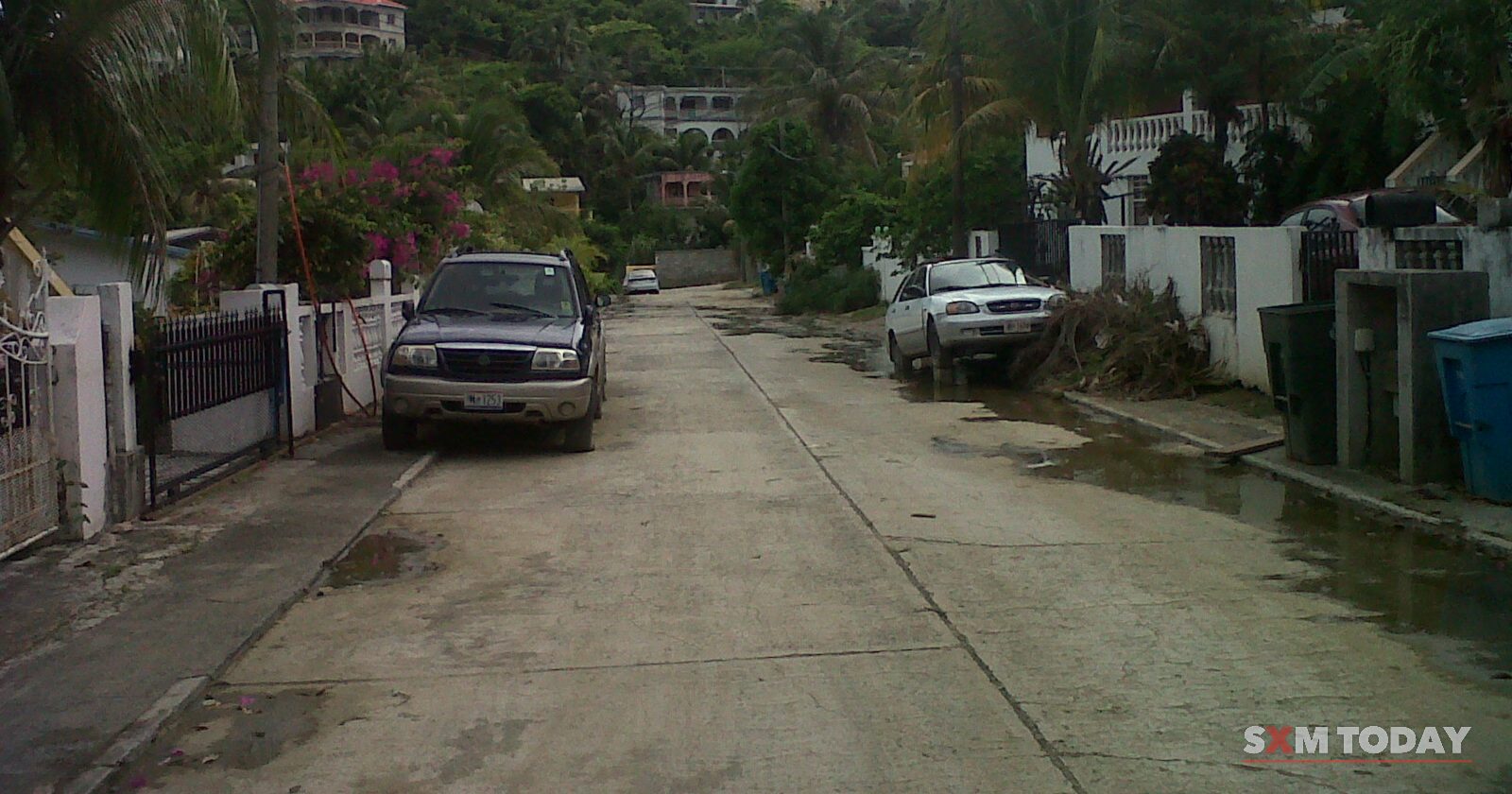 A concrete road with parked vehicles climbs between walls, trees and hillside houses under an overcast sky.