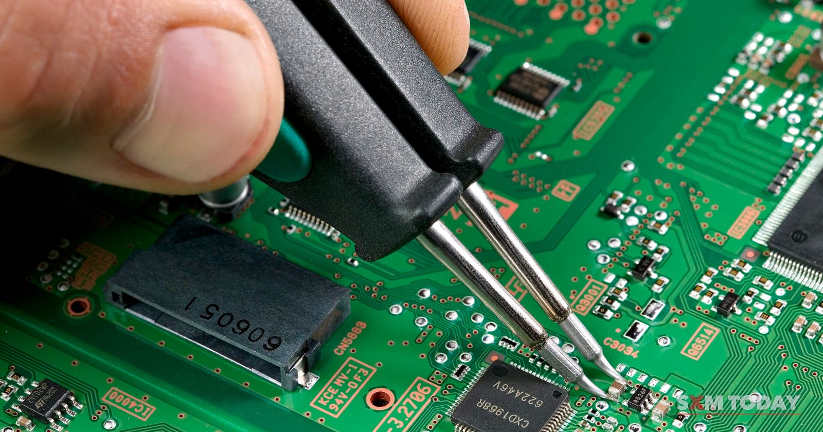 A hand works a soldering tool over a small component on a green circuit board.