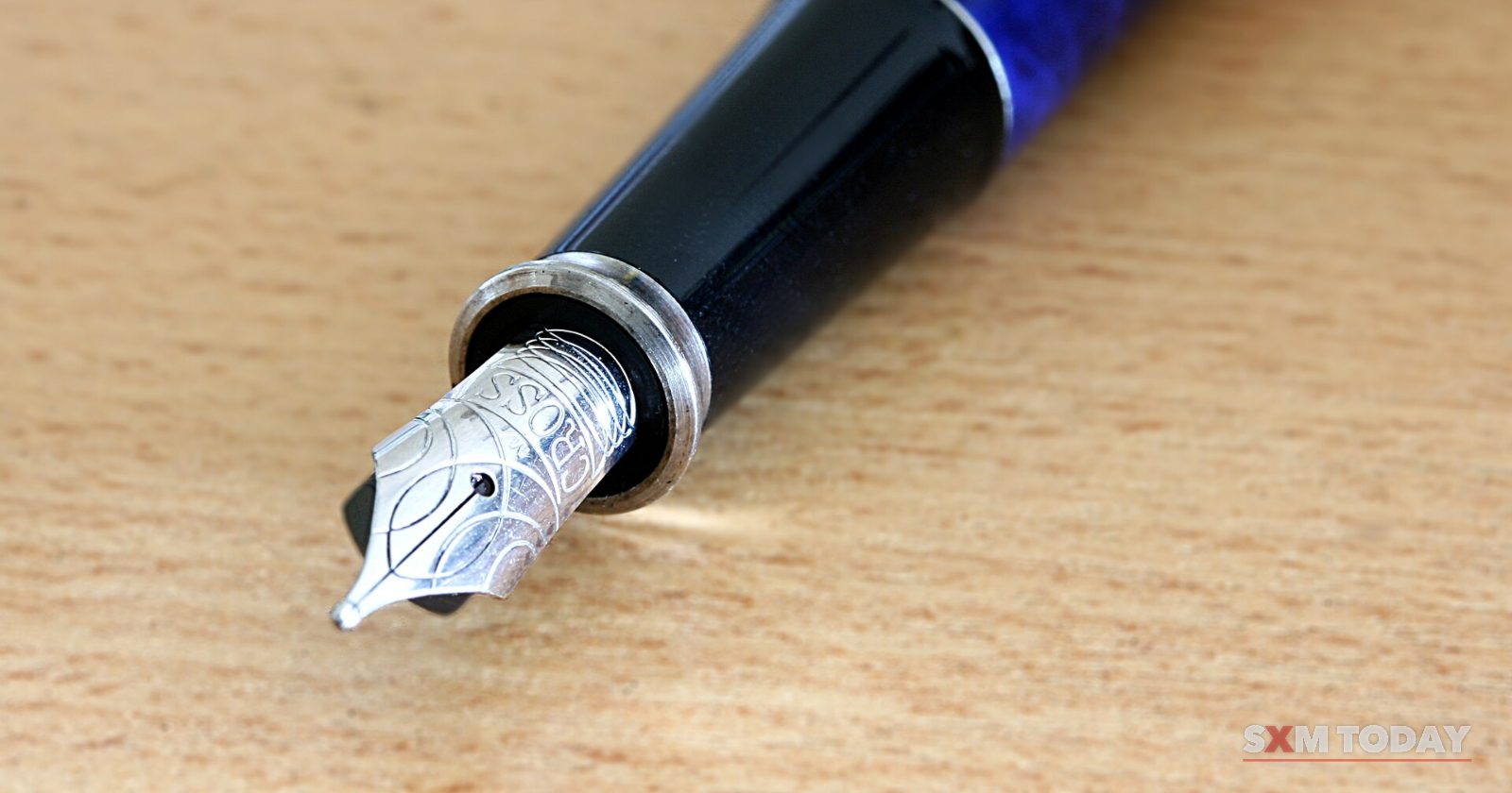Close-up of a fountain pen's silver nib and blue barrel lying on a light wooden desk.